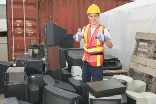 A young man wearing a yellow safety helmet, a high-visibility red and yellow vest, and purple gloves is standing outdoors at a waste collection site. He is smiling and giving a thumbs-up with both hands. In front of him is a pile of assorted discarded electronic devices and appliances, including old monitors, televisions, and computer components, all stacked on a paved surface. Behind him, there is a large red shipping container and a white tarpaulin covering part of the background, suggesting a commercial waste collection or disposal area typical of Shadwell. To the right, weathered wooden pallets are leaning against a wall or structure. The scene is well-lit, indicating daytime, and the focus is on the individual representing professional rubbish removal services provided by Commercial Waste Shadwell, emphasizing responsible disposal of electronic waste in the local area while capturing the landscape of a typical waste handling yard.
