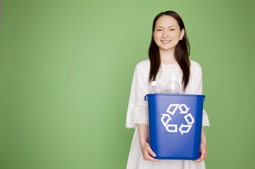 A young woman with long dark hair, smiling, stands against a plain light green background while holding a medium-sized blue recycling bin with a white recycling symbol on the front. She is dressed in a white, short-sleeved, loose-fitting top made of lightweight fabric. The bin appears to be made of plastic with a slightly textured surface, and it is filled with clear plastic bottles visible at the top. The woman’s posture is upright, and she is facing slightly to her left, with her hands gripping the sides of the bin at waist level. The setting has a clean, simple, professional appearance, suggesting a focus on eco-friendly waste management practices. This image is relevant for a rubbish and recycling service website such as commercialwasteshadwell.co.uk, particularly relating to environmentally conscious waste disposal or collection services in the Shadwell area of London, post code E1.