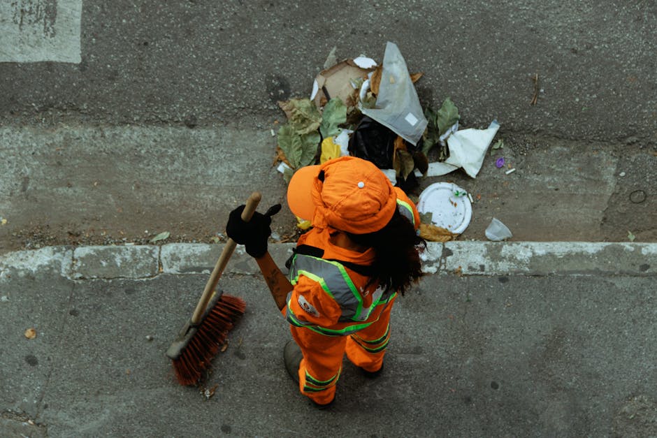 An individual wearing an orange high-visibility uniform and matching cap, holding a broom with a wooden handle and red bristles, is seen from above standing on a paved sidewalk near a concrete wall. T