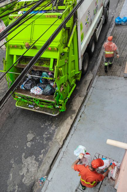 An individual wearing an orange high-visibility uniform and matching cap, holding a broom with a wooden handle and red bristles, is seen from above standing on a paved sidewalk near a concrete wall. T
